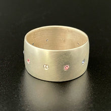 Load image into Gallery viewer, A wide sterling silver ring, with a line of flush set stones along the center. The stones are in the colors of the trans pride flag: light blue, pink, white, pink, and blue. All stones are lab grown: sapphires, rubies, and moissanite.