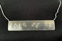 Load image into Gallery viewer, A necklace that reads "love is blind" in grade 2 braille. The piece is darkened, oxidized silver with polished silver dots, with the dots for the word "blind" done in gold.