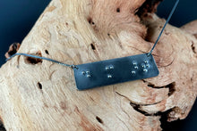 Load image into Gallery viewer, A functional, tactile braille necklace that reads "do more art" in grade 2 braille. The piece is dark, oxidized silver, draped over a piece of driftwood.
