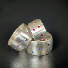 Load image into Gallery viewer, A stack of three rings - on the bottom is a thick silver ring with a scattering of flush set bright blue sapphires. Leaning against that is a similar ring with purple sapphires. Sitting on its side within the first ring is a smaller ring with a scattering of rubies.