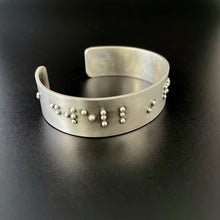 Load image into Gallery viewer, A sterling silver cuff on a black background, with accurately sized, spaced, and contracted braille. The portion seen at this angle reads "dwell in".