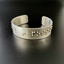 Load image into Gallery viewer, A sterling silver cuff bracelet, with accurately sized, spaced, and contracted braille. The portion that is visible reads "in possi".