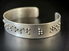 Load image into Gallery viewer, A sterling silver cuff on a black background. The cuff has accurately sized, spaced, and contracted braille. In this photo, braille reading "posed of nows" can be seen.