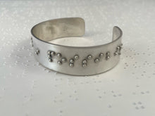 Load image into Gallery viewer, A sterling silver cuff bracelet resting on a brailled paper background. The cuff has accurately sized, spaced, and contracted braille, which in this photo reads "in possibil"