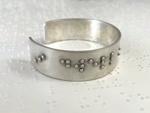 Load image into Gallery viewer, A sterling silver bracelet resting on brailled paper background. The bracelet has appropriately sized, spaced, and contracted braille, which in this photo reads "I dwell"
