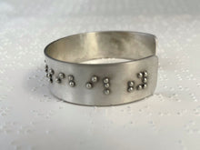 Load image into Gallery viewer, A sterling silver cuff bracelet resting on a background of brailled paper. The cuff has accurately sized, spaced, and contracted braille, which in this photograph reads, "ssibility"