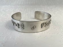Load image into Gallery viewer, A sterling silver cuff bracelet resting on a background of brailled paper. The bracelet has appropriately sized, spaced, and contracted braille. The portion visible in this photograph reads "dwell in poss"