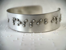 Load image into Gallery viewer, A close up on a sterling silver cuff bracelet, which is resting on some brailled paper. The bracelet has accurately sized, spaced, and contracted braille on it. The portion of the braille visible in this photograph reads, "possibil"