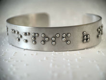 Load image into Gallery viewer, A sterling silver cuff on a background of brailled paper. The cuff has accurately sized, spaced, and contracted braille. In this photo, braille reading "composed of" can be seen.