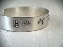 Load image into Gallery viewer, A sterling silver cuff on a background of brailled paper. The cuff has accurately sized, spaced, and contracted braille. In this photo, braille reading "forever is" can be seen.