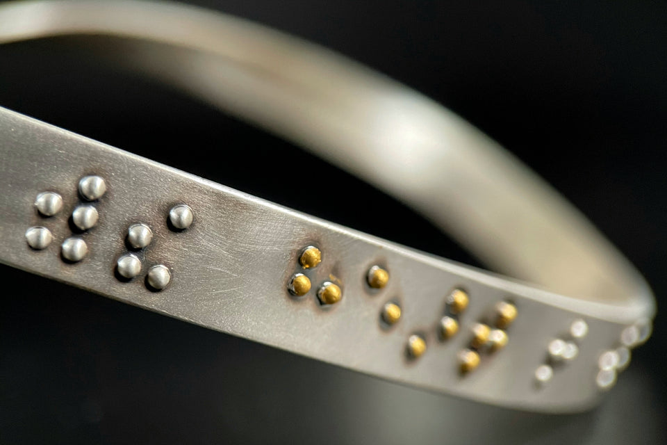 A closeup of a silver neckpiece with braille bumps. The visible braille reads "with the heart" and the word "heart" is done in gold.