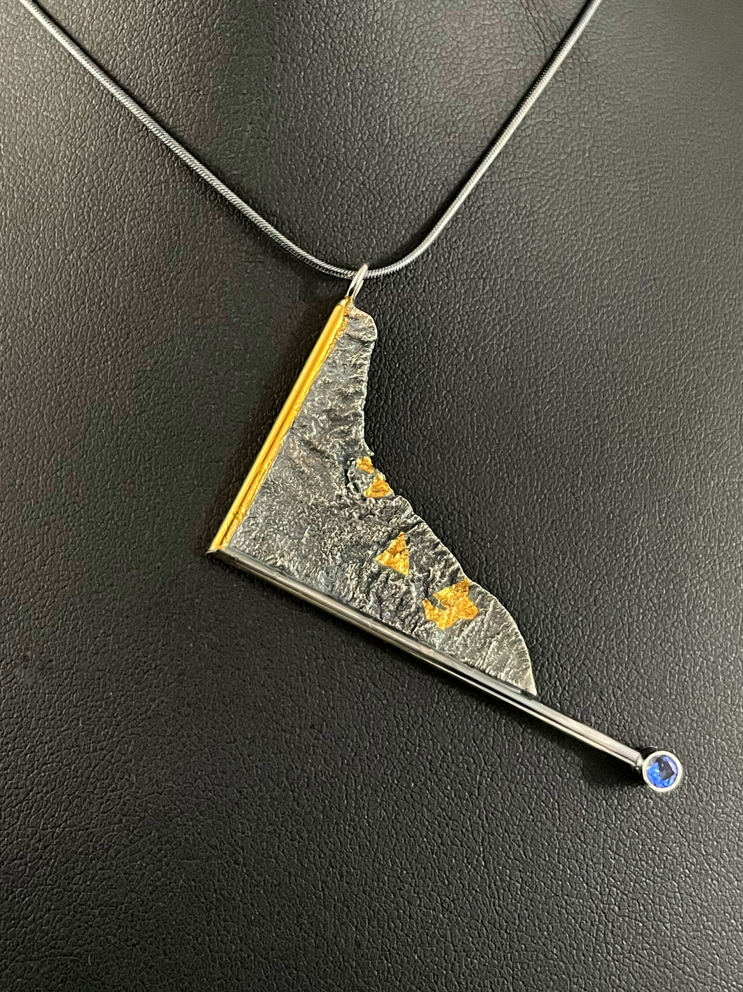 A triangular pendant, hanging askew. It is framed on two sides, one in gold covered wire, and one in silver. At the end of the framing silver wire is a lab grown blue sapphire. The body of the pendant is reticulated silver, with a rough, organic texture. Small pools of gold are hidden in the crevices.