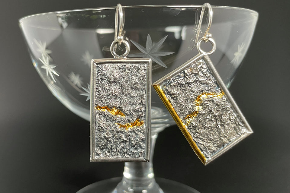Two rectangular silver earrings with deep reticulated texture framed in silver wire. Both have gold tracing a valley across each piece. The earring on the right hangs at an angle, with one part of the silver frame done in gold.