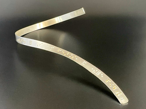 A sinuous braille neckpiece made of sterling silver. The braille dots along it read, 