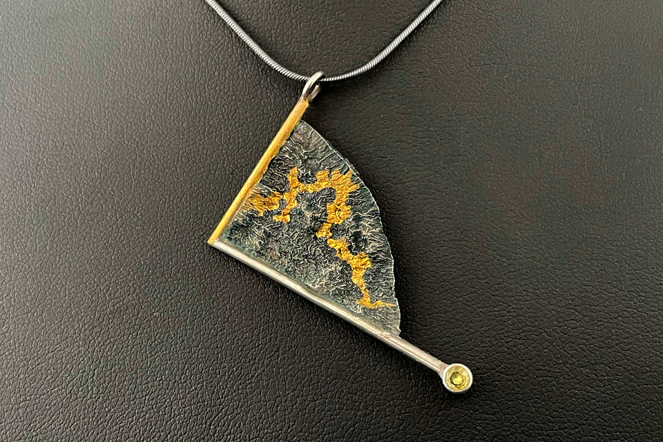 A triangle pendant, hanging askwe. It is framed on one side in gold and another in silver wire. The silver frame overshoots the body of the pendant and ends in a lab grown yellow sapphire. The main body of the pendant is a darkened reticulated silver in a rough organic texture, with is a winding path of gold running through the crevices of the silver.