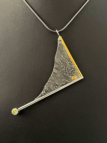 A triangular pendant hanging askew. It is framed on two sides with wire, one gold and one silver. The end of the silver frame runs off past the body of the pendant and terminates in a lab-grown yellow sapphire. The body of the pendant is reticulated silver, in a rough organic texture. A small spot of gold is nestled in the valley of the silver.