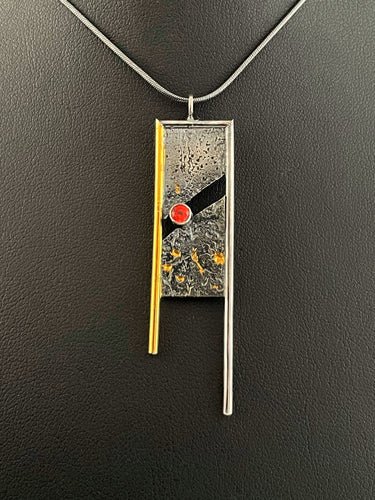 A rectangular pendant, framed on three sides with wire. The wire on the left is gold, with the top and right sides being silver. The frames hang down past the body of the pendant on the bottom. The main pendant is reticulated silver, with a rough organic texture and flecks of gold in the crevices. It is split diagonally through the middle, with a lab grown orange sapphire set in the gap.