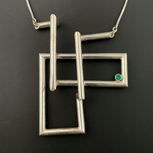 A twist of silver tubing set at right angles, overlapping itself in a double L shape. In the right-hand corner is a lab grown bright green emerald.