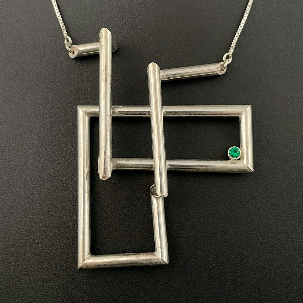 A twist of silver tubing set at right angles, overlapping itself in a double L shape. In the right-hand corner is a lab grown bright green emerald.