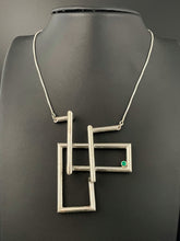 Load image into Gallery viewer, A twist of silver tubing set at right angles, overlapping itself in a double L shape. In the right-hand corner is a lab grown bright green emerald. Piece is approximately 3 inches tall and 2 inches wide.
