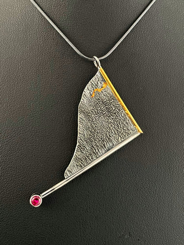 A triangular pendant, hanging askew. It is framed on two sides, one in gold covered wire, and one in silver. At the end of the framing silver wire is a lab grown ruby. The body of the pendant is reticulated silver, with a rough, organic texture. A single small stripe of gold runs down a crevice.