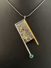 Load image into Gallery viewer, A rectangular pendant, framed on three sides with wire and hanging askew. The wire on the left is gold, with the top and right sides being silver. The frames hang down past the body of the pendant on the bottom. Off of the end of the silver frame is a lab-grown emerald. The main pendant is reticulated silver, with a rough organic texture and a river of Gold winding through the valley of the silver. 
