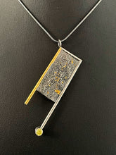 Load image into Gallery viewer, A rectangular pendant, framed on three sides with wire and hanging askew. The wire on the left is gold, with the top and right sides being silver. The frames hang down past the body of the pendant on the bottom. Off of the end of the silver frame is a lab-grown yellow sapphire. The main pendant is reticulated silver, with a rough organic texture and flakes of gold hidden amongst the crevices of the metal.
