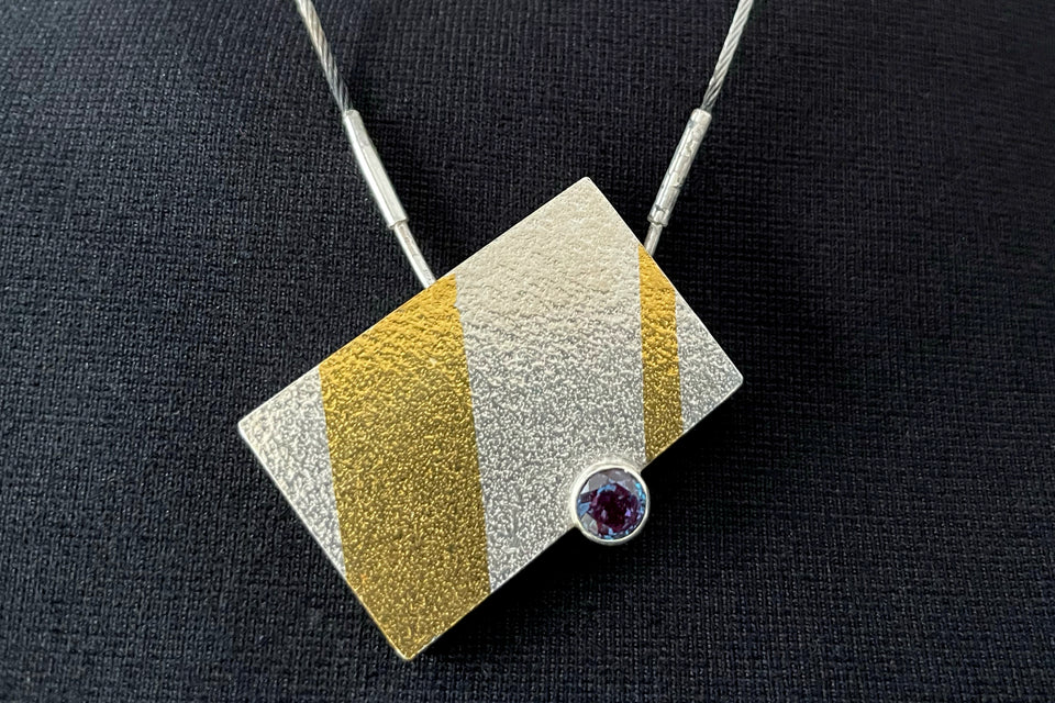 A rectangular necklace of textured silver, hanging at an angle. Two stripes of gold run almost vertically: a thick one on the left side, and thin on the right. Between them at the bottom is a tube-set round blue/purple alexandrite gemstone.