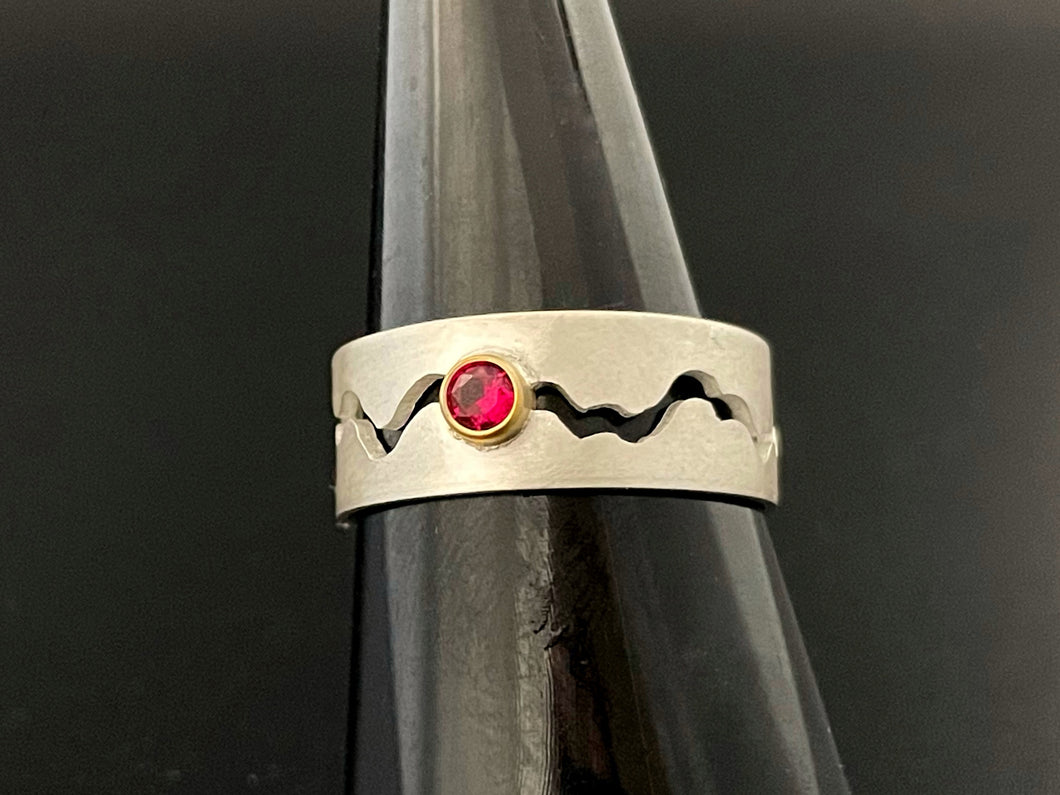 A sterling silver ring with a winding gap running down the middle of the band. There is a lab-grown round ruby in an 18k yellow gold setting in the middle of the gap.