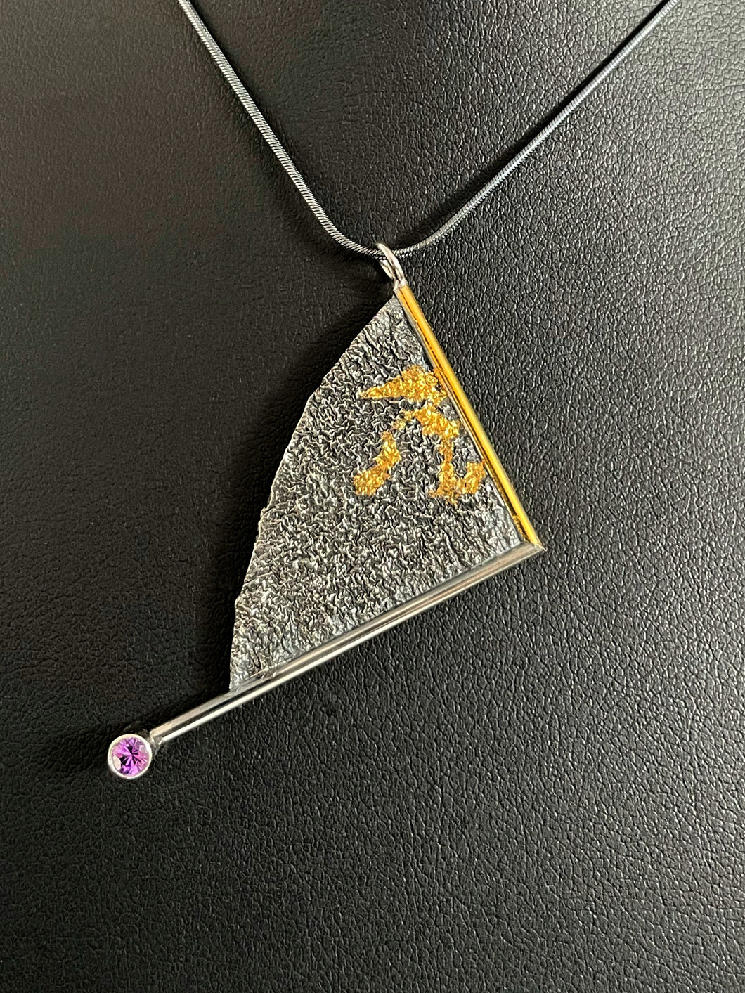 A triangular pendant, hanging askew. It is framed on two sides, one in gold covered wire, and one in silver. At the end of the framing silver wire is a lab grown purple sapphire. The body of the pendant is reticulated silver, with a rough, organic texture. Small wandering strips of gold are nestled in the upper half.