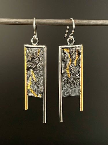 Long rectangular earrings. They are framed on the top and inside edge in silver wire, and on the outside edge in 24k gold. Each leg of the frame hangs down below the bottom of the body, with the inside silver leg longer. The body of the earring is a rough, wild texture reminiscent of land seen from air. It's oxidized and dark, with the high parts polished, and trails of gold running in the valleys of the texture.