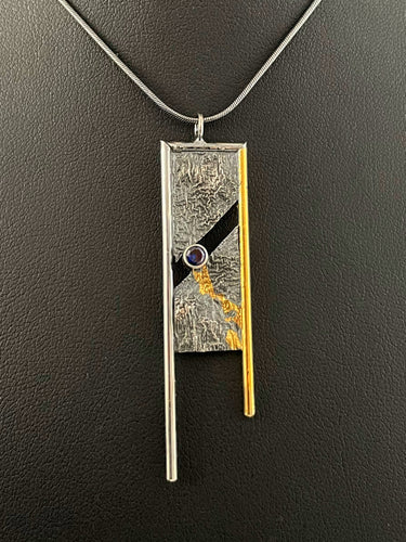 A rectangular pendant, framed on three sides with wire. The wire on the left is gold, with the top and right sides being silver. The frames hang down past the body of the pendant on the bottom. The main pendant is reticulated silver, with a rough organic texture. It is split diagonally through the middle, with a lab grown blue sapphire set in the gap. A path of gold leads down from the gemstone through the crevices in the silver.