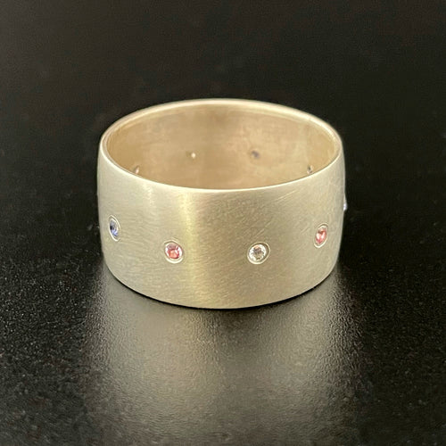 A wide sterling silver ring, with a line of flush set stones along the center. The stones are in the colors of the trans pride flag: light blue, pink, white, pink, and blue. All stones are lab grown: sapphires, rubies, and moissanite.