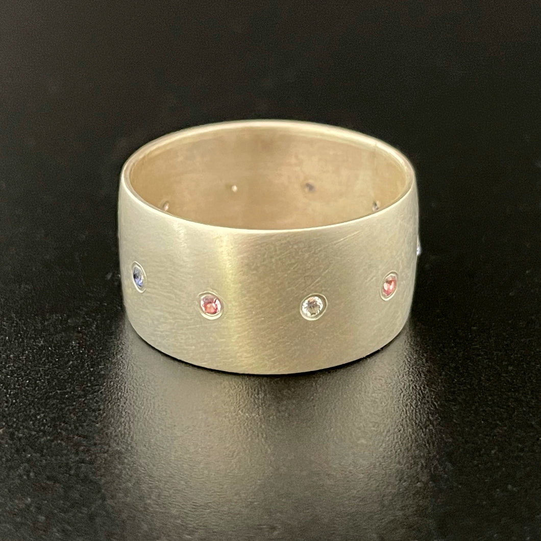 A wide sterling silver ring, with a line of flush set stones along the center. The stones are in the colors of the trans pride flag: light blue, pink, white, pink, and blue. All stones are lab grown: sapphires, rubies, and moissanite.