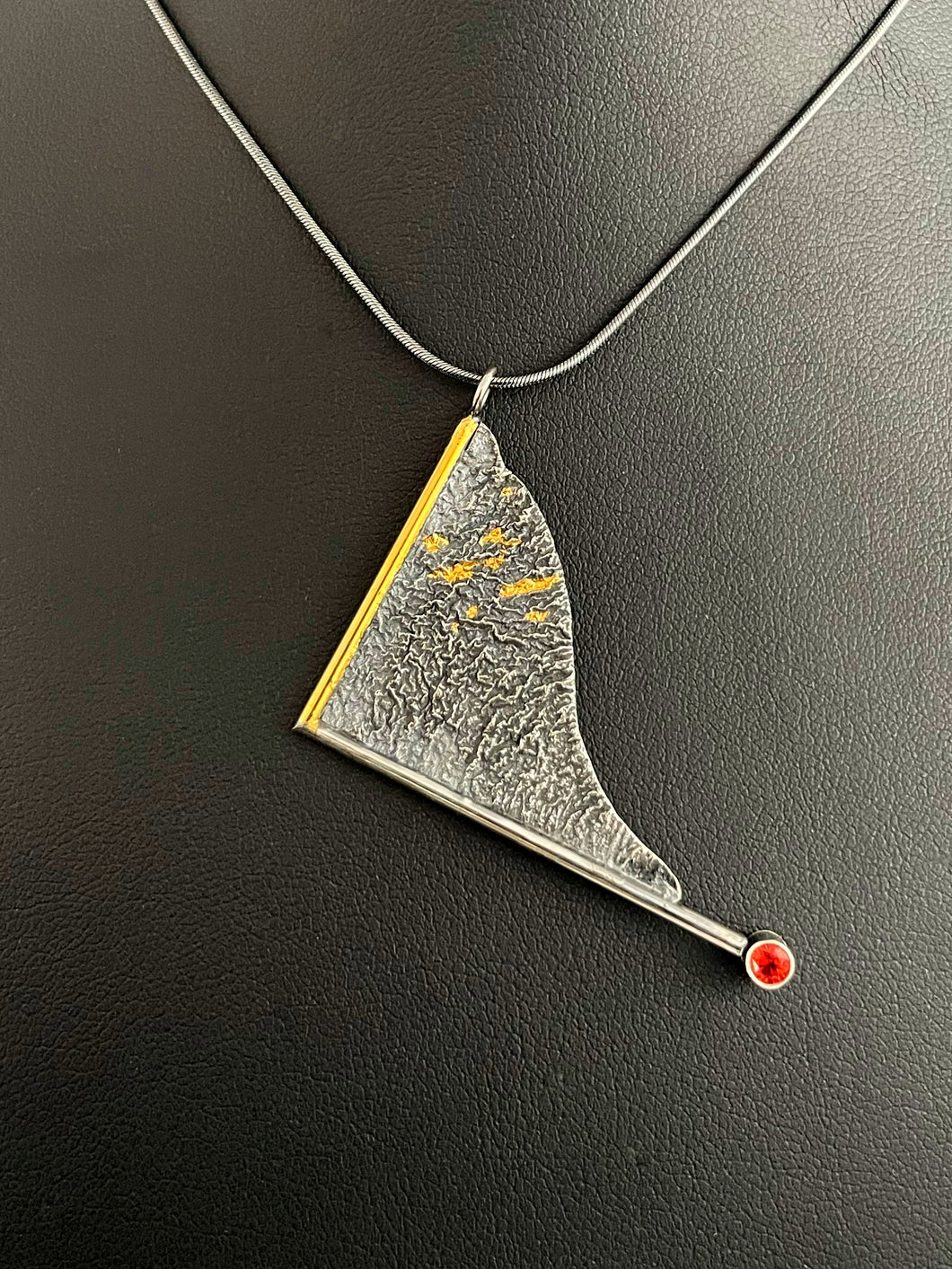 A triangular pendant, hanging askew. It is framed on two sides, one in gold covered wire, and one in silver. At the end of the framing silver wire is a lab grown orange sapphire. The body of the pendant is reticulated silver, with a rough, organic texture. Small stripes of gold are nestled in the upper corner.