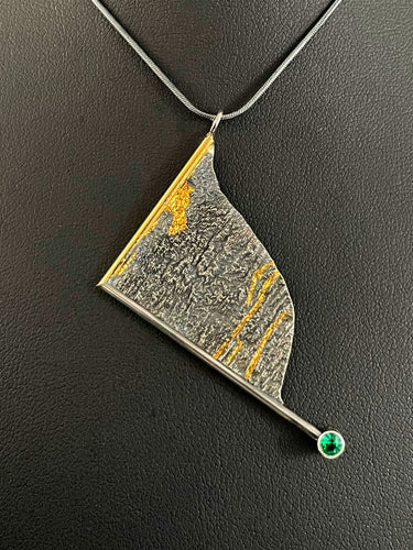 A triangular pendant, hanging askew. It is framed on two sides, one in gold covered wire, and one in silver. At the end of the framing silver wire is a lab grown emerald. The body of the pendant is reticulated silver, with a rough, organic texture. Small stripes of gold run along the crevices.