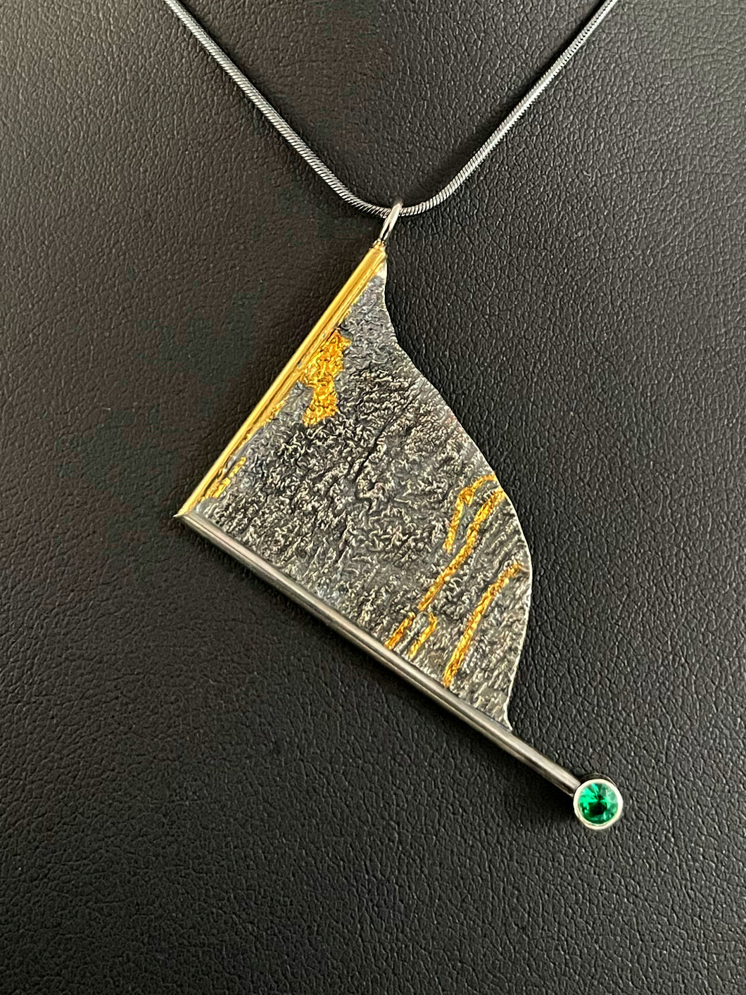 A triangular pendant, hanging askew. It is framed on two sides, one in gold covered wire, and one in silver. At the end of the framing silver wire is a lab grown emerald. The body of the pendant is reticulated silver, with a rough, organic texture. Small stripes of gold run along the crevices.