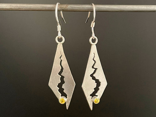 A pair of long diamond shaped sterling silver earrings. There's a long winding gap cut into the silver, which gets wider towards the bottom and culminates in a round, lab-grown yellow sapphire.