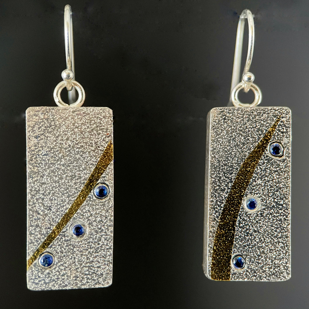 Rectangular silver earrings. This side of them is textured silver with similar but not identical gold arcs. Below the arcs there is a line of 3 lab-grown sapphires that match the flow of the gold.