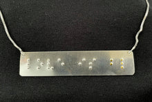 Load image into Gallery viewer, A necklace that reads &quot;love is blind&quot; in grade 2 braille. The piece is darkened, oxidized silver with polished silver dots, with the dots for the word &quot;blind&quot; done in gold.
