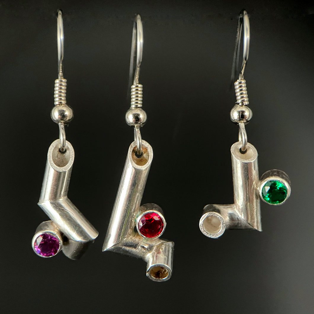 Three earrings hanging side by side. Each is made of sterling silver tubing with sharp bends making it twist and turn in three dimensions. Each has a different gemstone sitting in one of the bends - purple sapphire, ruby, and emerald.