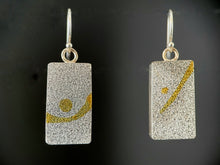 Load image into Gallery viewer, Textured silver rectangular earrings, each with similar but not identical gold patterns on them. The earring on the left has a sharply slanted gold line with a dot above it, while the earring on the right has a curved gold line and a larger dot nestled within the curve.
