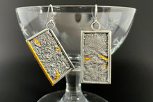 Load image into Gallery viewer, A pair of reversible earrings. These are rectangles framed in silver wire. The one on the left hangs at an angle. Within the frames is rough-textured reticulated silver. In some of the valleys of the silver texture are small triangles of bright gold. On the tilted piece, on piece of the silver wire frame is also gold.
