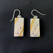 Load image into Gallery viewer, Rectangular silver earrings. This side of them is textured silver with a lined patter. Following those diagonal lines are two gold stripe. Between them are 2 purple sapphires on one earring and 3 on the other, following the angle of the gold.

