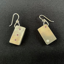 Load image into Gallery viewer, These rectangular silver earrings hang at an angle. The earring on the left has a purple sapphire in the upper right corner and a larger sparklly moissanite in the lower left. The earring on the right has three purple sapphires in a vertical line down the center.
