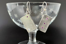 Load image into Gallery viewer, These rectangular silver earrings hang at an angle. The earring on the left has a purple sapphire in the upper right corner and a larger sparklly moissanite in the lower left. The earring on the right has three purple sapphires in a vertical line down the center.
