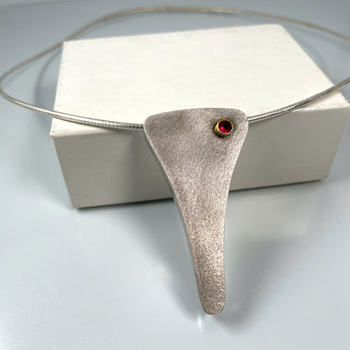 A triangular shaped necklace. The pendant has gentle curves making a vague Y shape before meeting in a curved, rounded point. The piece is textured sterling silver, slightly darkened. In the upper right corner of the piece is a round, brilliant cut lab-grown ruby, in a gold foiled tube bezel.