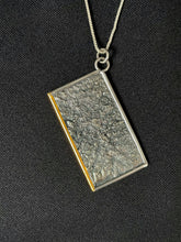 Load image into Gallery viewer, A rectangular silver necklace, hanging at an angle. The piece is roughly textured reticulated silver that has the low points darkened. There is a sort of crater shape in the upper part, which has been highlighted by 3 lines in gold, shaped kind of like an hourglass. The bottom side of the frame is done in gold.
