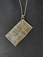 Load image into Gallery viewer, A rectangular silver necklace, hanging at an angle. The piece is roughly textured reticulated silver with a wire framing. There is a sort of crater shape in the upper part, which has been highlighted by 3 lines in gold, shaped kind of like an hourglass. The bottom side of the frame is done in gold.
