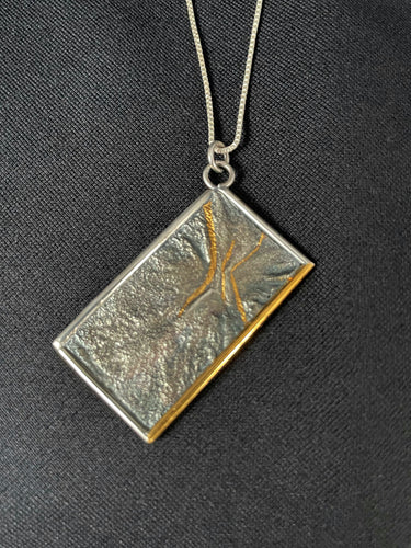 A rectangular silver necklace, hanging at an angle. The piece is roughly textured reticulated silver with a wire framing. There is a sort of crater shape in the upper part, which has been highlighted by 3 lines in gold, shaped kind of like an hourglass. The bottom side of the frame is done in gold.